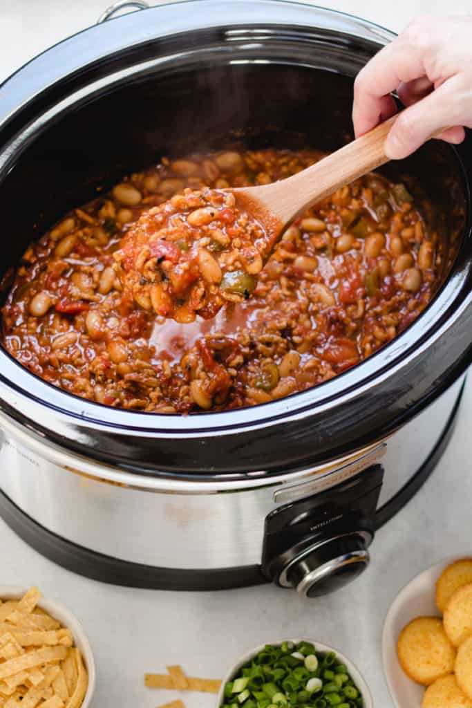 A slow cooker with chipotle chicken chili inside, a hand holding a wooden spoonful out of the chili.