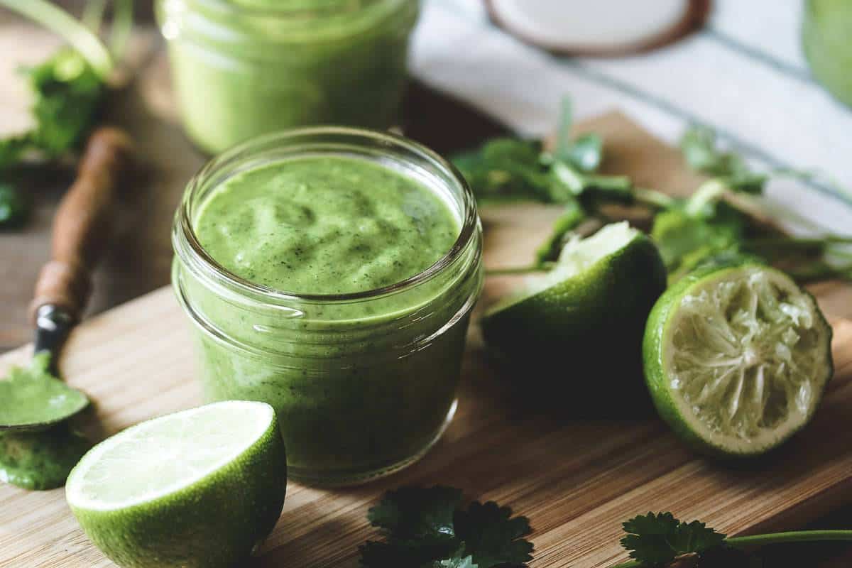 A jar of cilantro vinagraitte surrounded by limes and cilantro on a small bamboo cutting board.