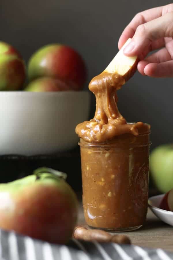 A hand dipping into Peanut Butter Caramel Apple Dip that is spilling over the side of a mason jar. Red and green apples.