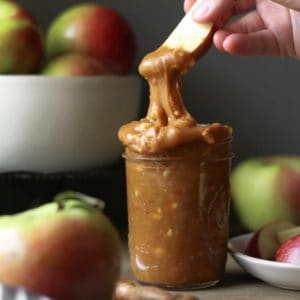 A hand dipping into Peanut Butter Caramel Apple Dip that is spilling over the side of a mason jar. Red and green apples.