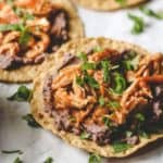 three tostadas with refried black beans and chicken tinga on parchment paper, shredded chicken in tinga sauce, sauce, tostadas, and refried black beans