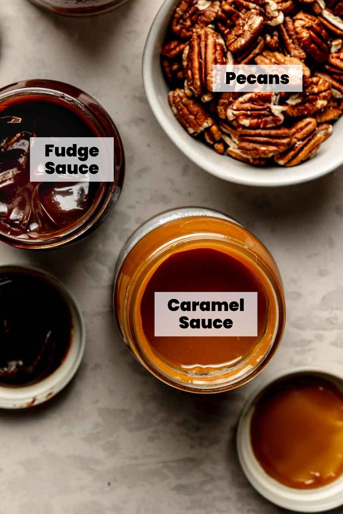 Open jars of fudge sauce and caramel sauce, next to a bowl of pecans.