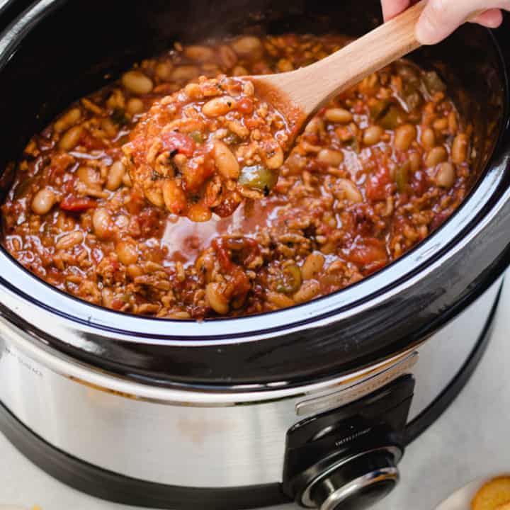 A slow cooker with chipotle chicken chili inside, a hand holding a wooden spoonful out of the chili.