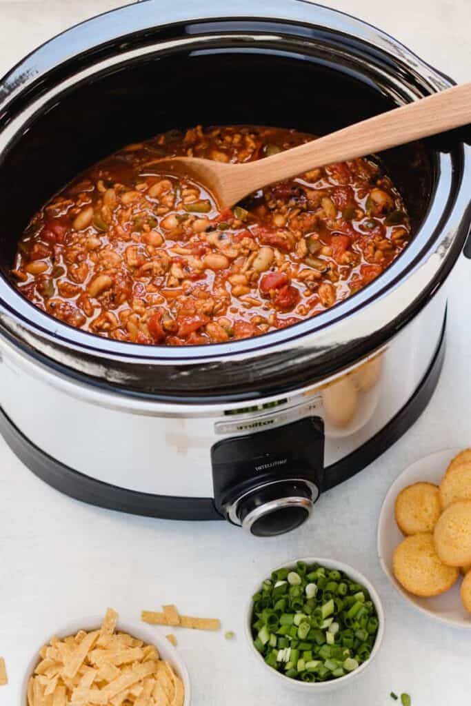 A slow cooker with chipotle chicken chili inside, a wooden spoon resting on edge.