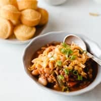 a bowl of chili with cheddar cheese and green onions on top. Mini cornbread muffins in background.