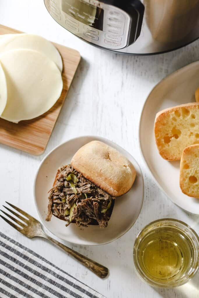 An Italian beef sandwich on a white plate near the instant pot, cheese and extra rolls on the side