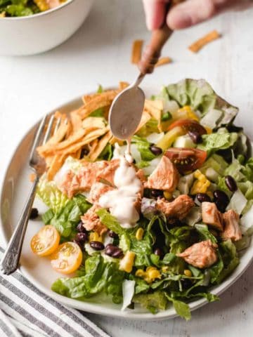 Pouring chipotle ranch dressing over a southwest bbq chicken salad piled high on a plate.