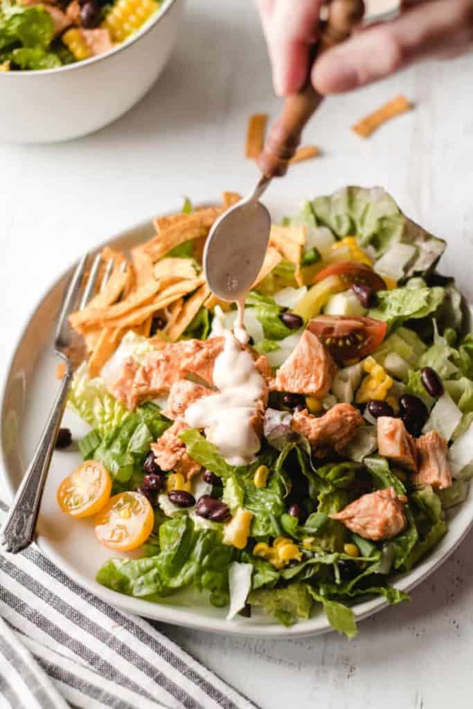 Pouring chipotle ranch dressing over a southwest bbq chicken salad piled high on a plate.