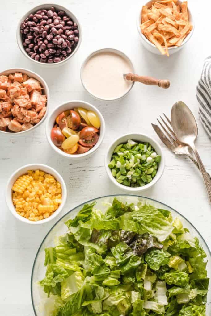 Salad ingredients laid out in bowls, beans, corn, lettuce, chips, chicken, and salad dressing.