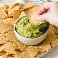 Dipping a tortilla chip into a bowl of easy guacamole surrounded by chips.