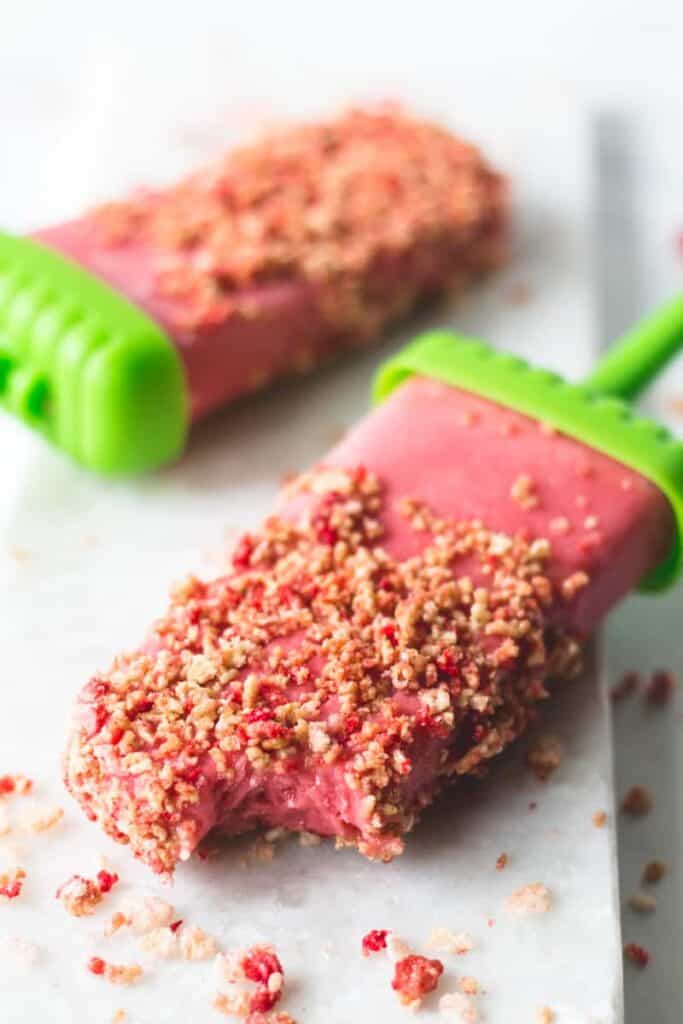 Two strawberry Shortcake Popsicles on marble slab, one with a bite taken from it.