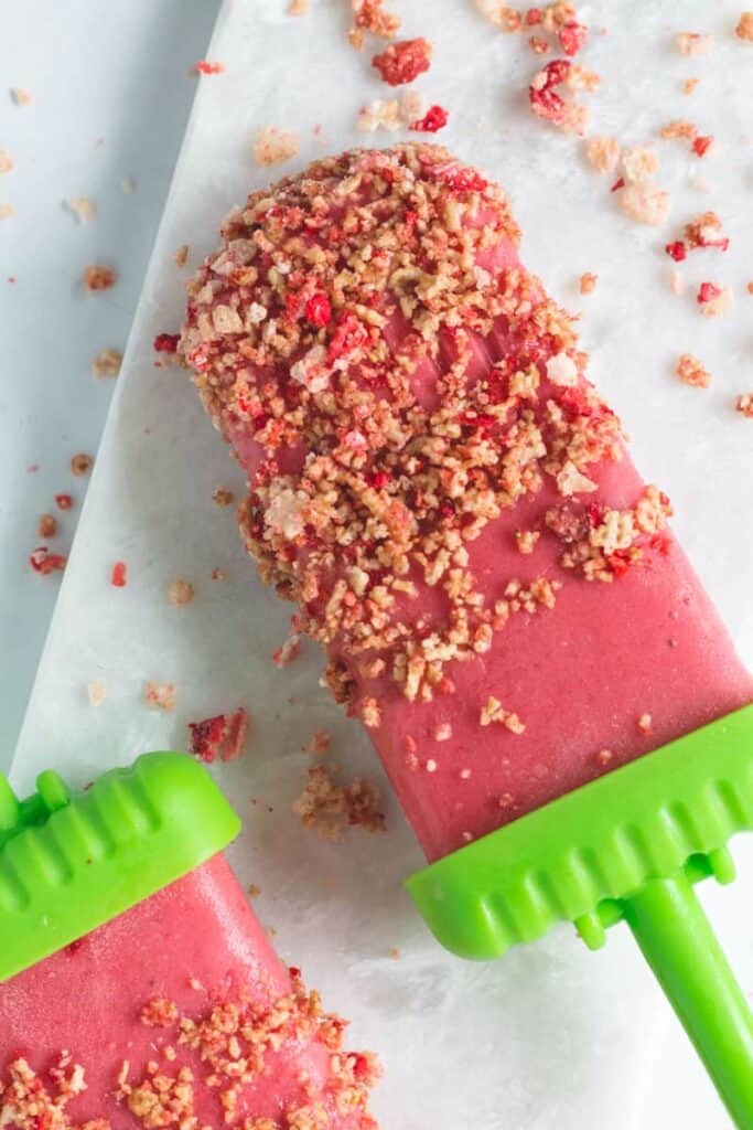 A bright pink strawberry shortcake popsicle covered in cinnamon Chex and strawberry crunch with a green popsicle handle.  