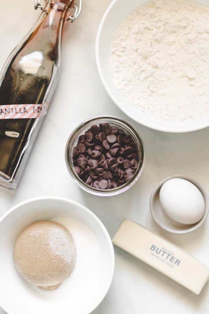 Ingredients laid flat for chocolate chip cookies, egg, butter, flour, sugars, chocolate chips, and a bottle of vanilla.