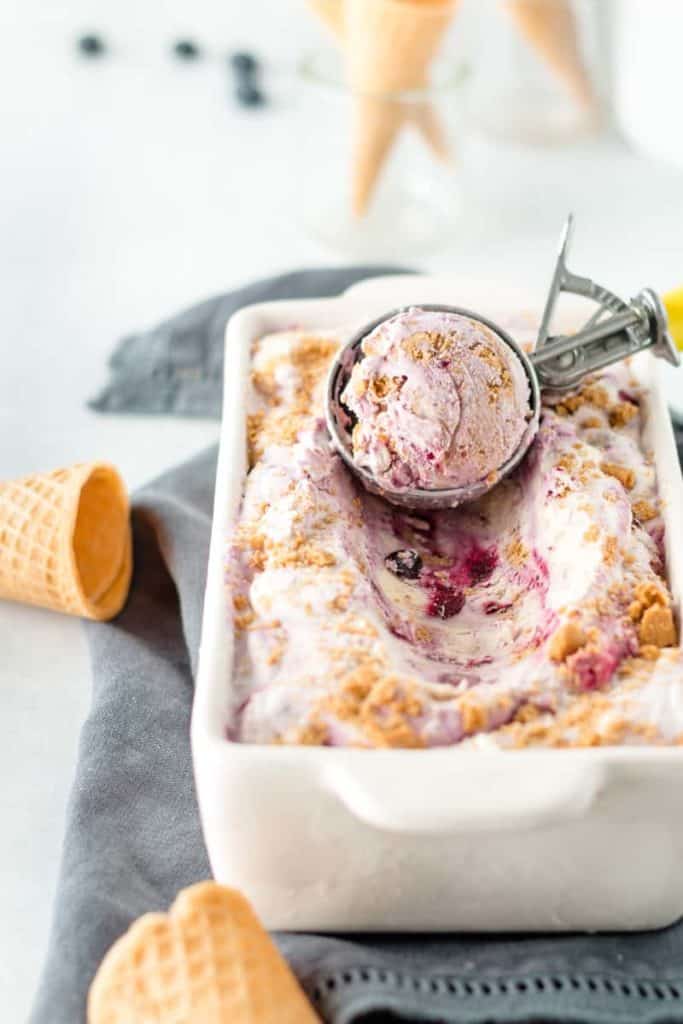 Blueberry cheesecake no churn ice cream in a loaf pan with a scoop in an ice cream scoop.  Sugar cones to the side.