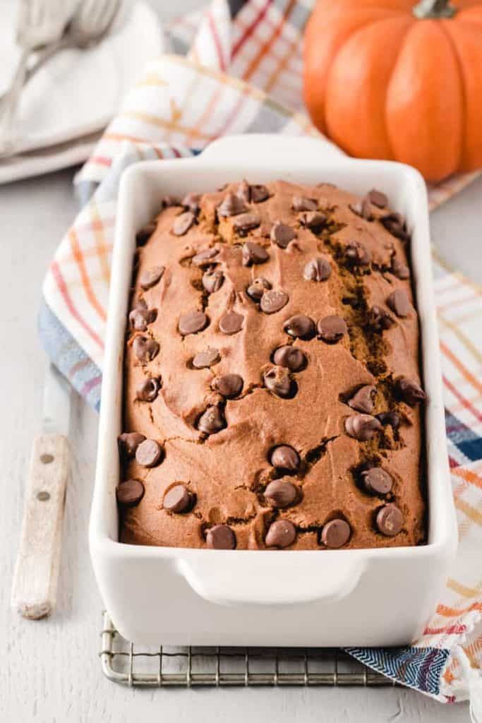A loaf of gluten-free chocolate chip pumpkin bread in a white loaf pan on a plaid towel. A pumpkin in background.