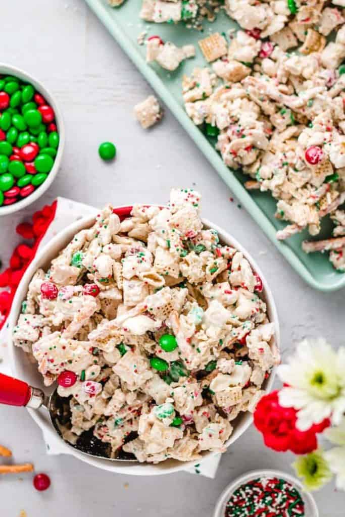 Christmas chex mix in a bowl next to m&m's and more on a cookie sheet.