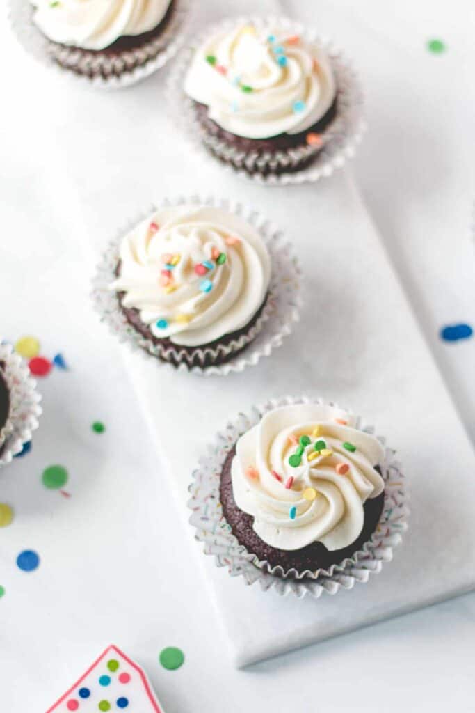 Gluten-free chocolate cupcakes with vanilla frosting and rainbow sprinkles on a marble platter.