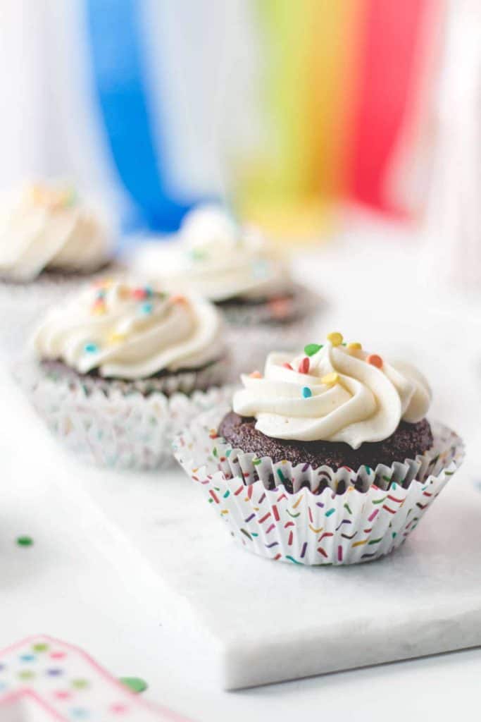Gluten-free chocolate cupcakes with vanilla frosting and rainbow sprinkles. Rainbow streamers in the background.