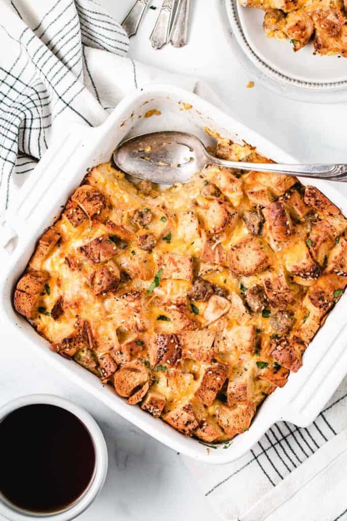 Gluten-free Everything Bagel Strata in a white square baking dish. It is brown and crispy on top with a scoop removed and spoon resting to the side.