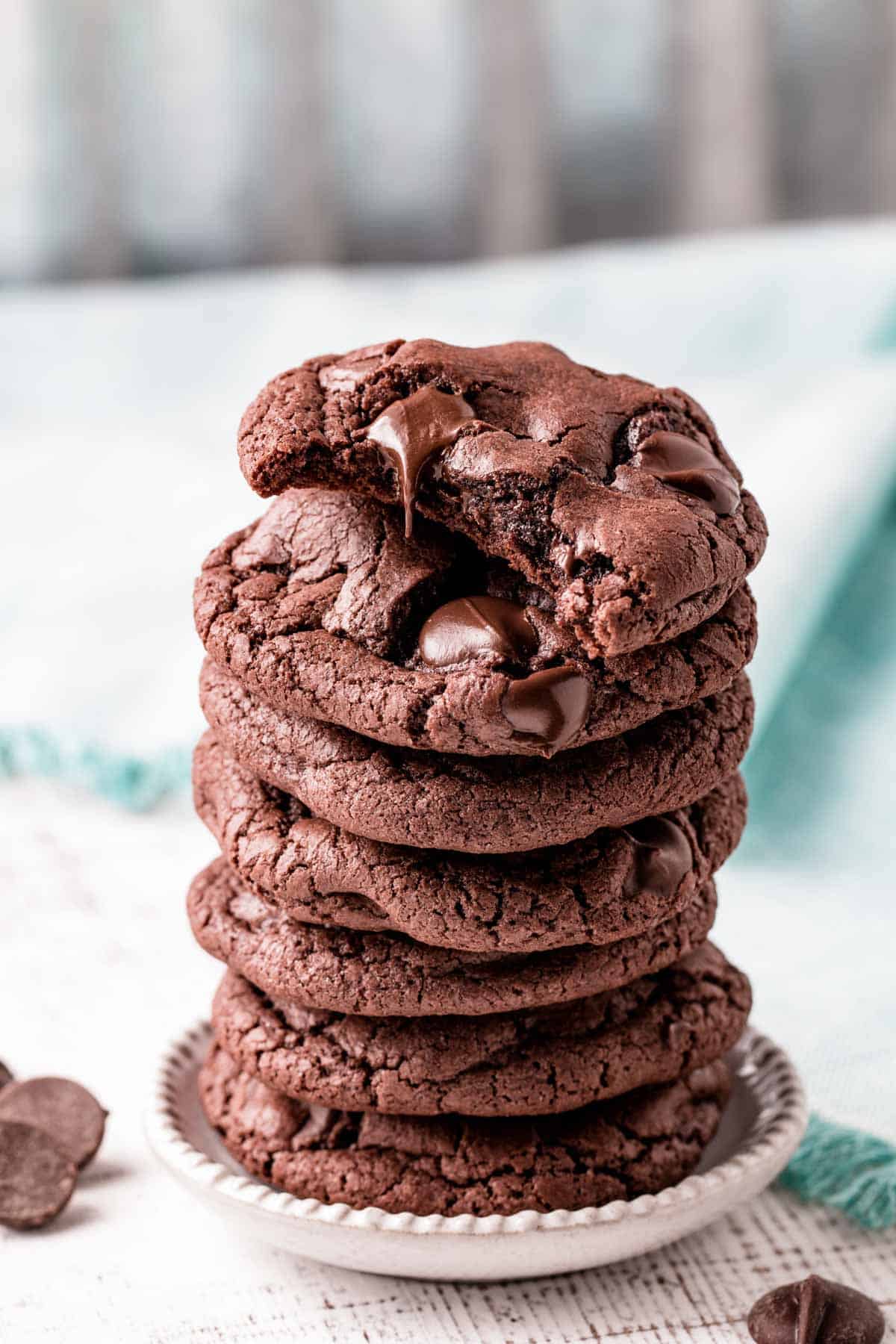 A stack of gluten-free double chocolate cookies.  The top cookie is half eaten. There is a chocolate drip falling from one of the chocolate chips.