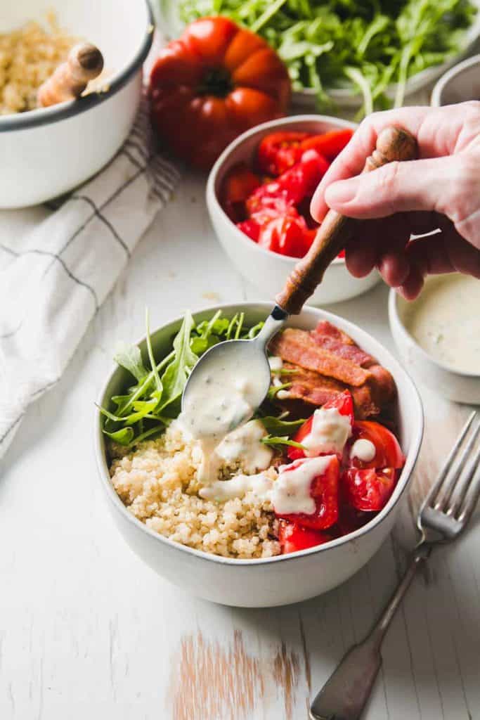 A bowl of quinoa, bacon, red tomatoes, and arugula is drizzled with yogurt dressing.