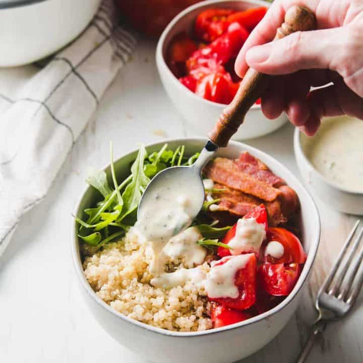 A bowl of quinoa, bacon, red tomatoes, and arugula is drizzled with yogurt dressing.
