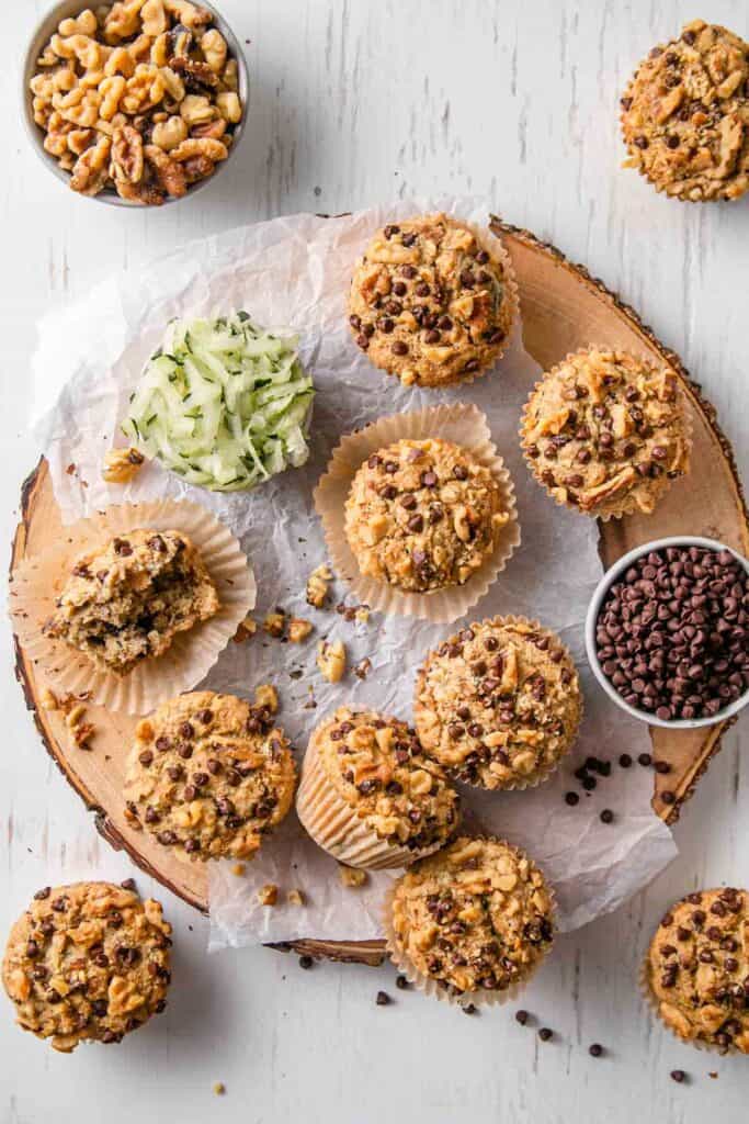 Gluten-free zucchini muffins on a round wood slice platter next to shredded zucchini and a bowl of chocolate chips.