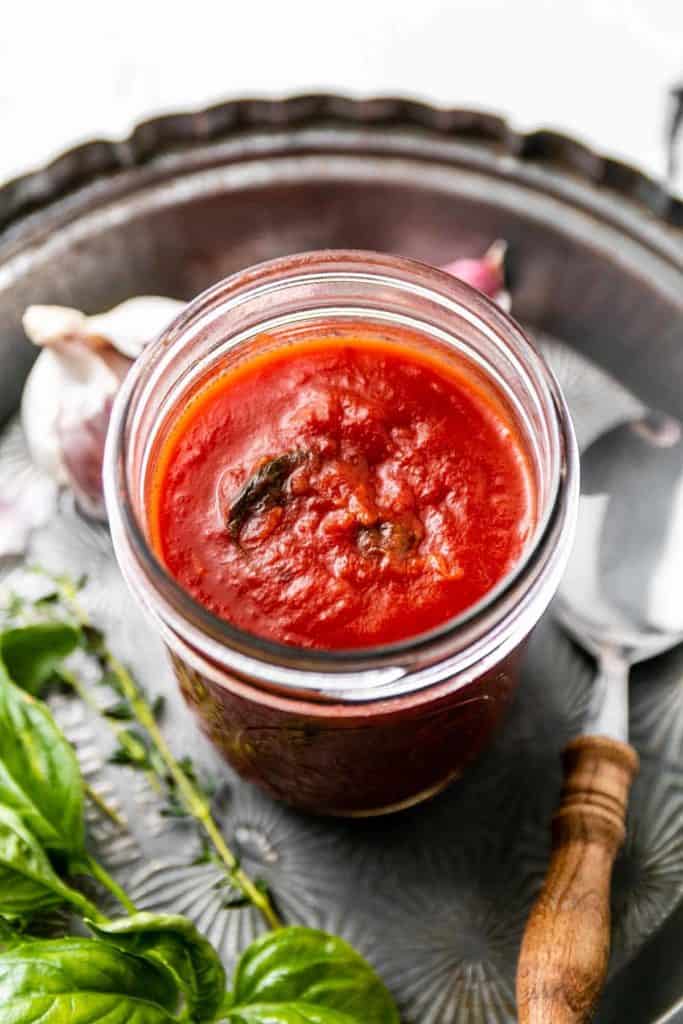 Pizza sauce with wilted basil leaves in a mason jar. On old oven pie dish surrounded by fresh herbs, garlic and an old spoon.