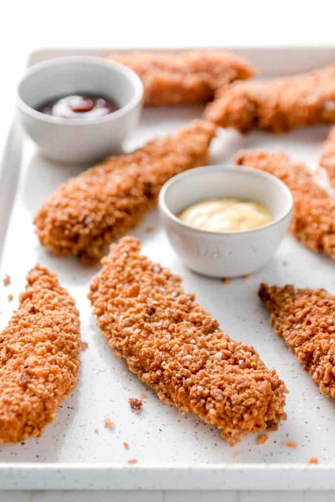 A tray of golden chicken tenders next to small cups of bbq sauce and honey mustard.