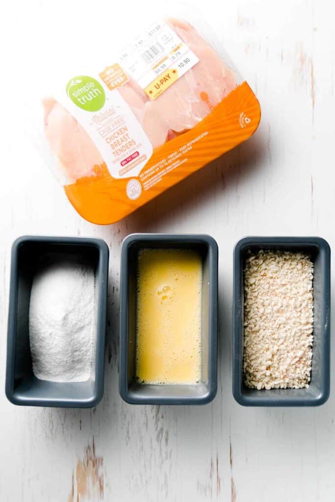 Chicken tenders in the package next to three mini loaf pans with rice flour, egg mixture, and breadcrumbs.