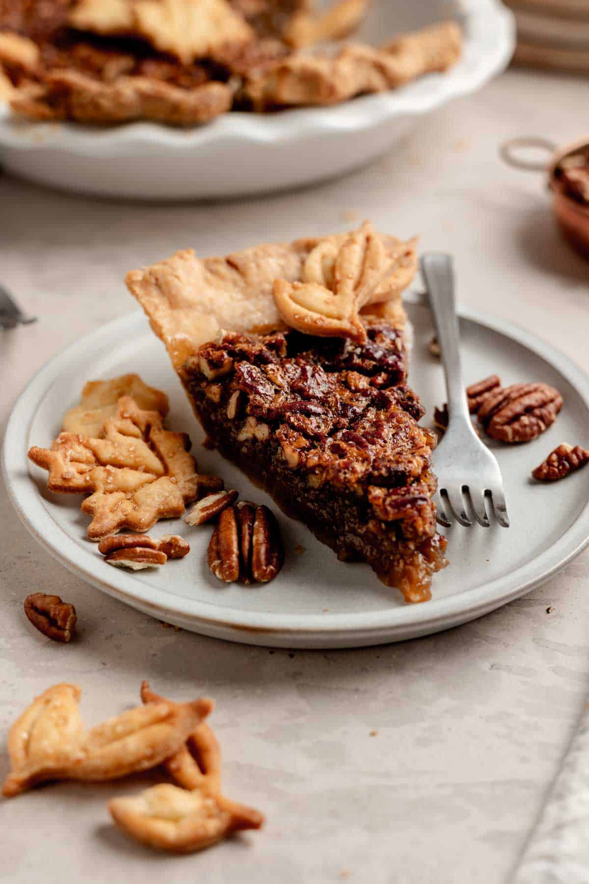 A slice of gluten-free pecan pie on a small plate next to pecans and leaf-shaped pie crust cut outs.