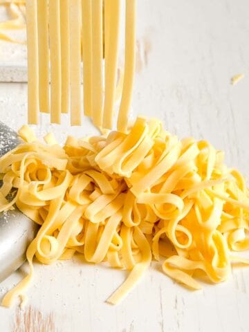 Fresh gluten free egg noodles in a pile, some dangling from pasta cutter above.