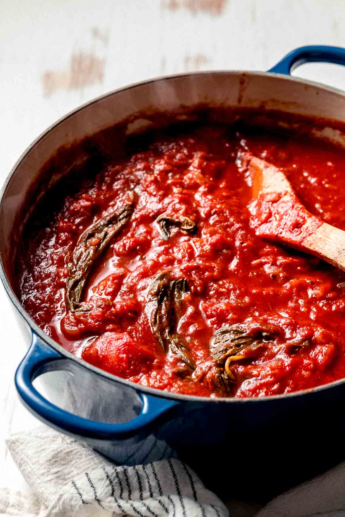 A large blue le crueset pot full of gluten-free tomato sauce. Wilted basil and a wooden spoon rest on top.