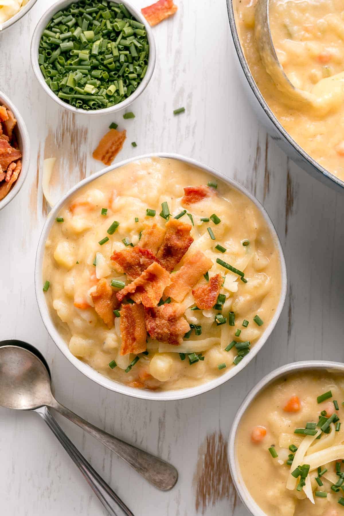 Sopa de papa sin gluten en un tazón de crema adornada con cebollino y tocino al lado de una sartén.