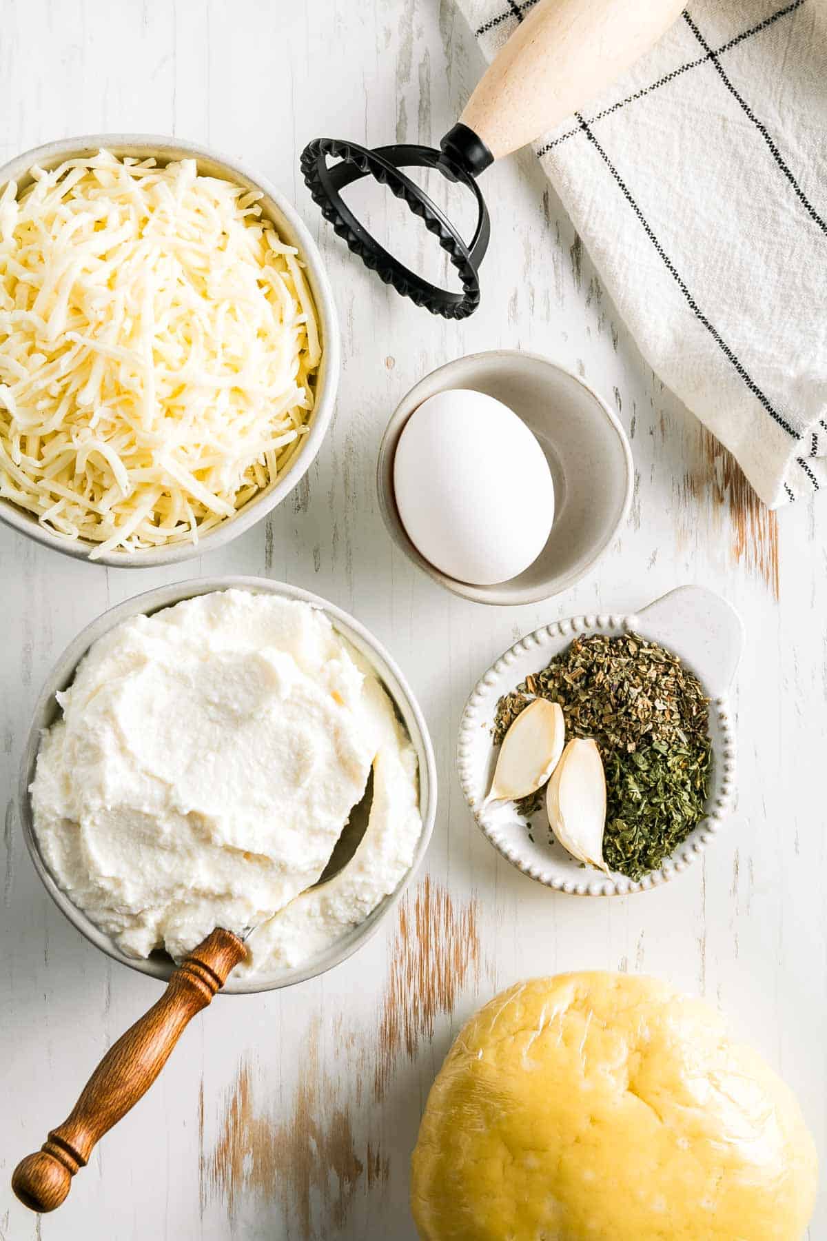 Pasta dough wrapped in plastic next to a bowl of ricotta cheese and shredded Italian blend cheese along with an egg, spices and a ravioli stamp.