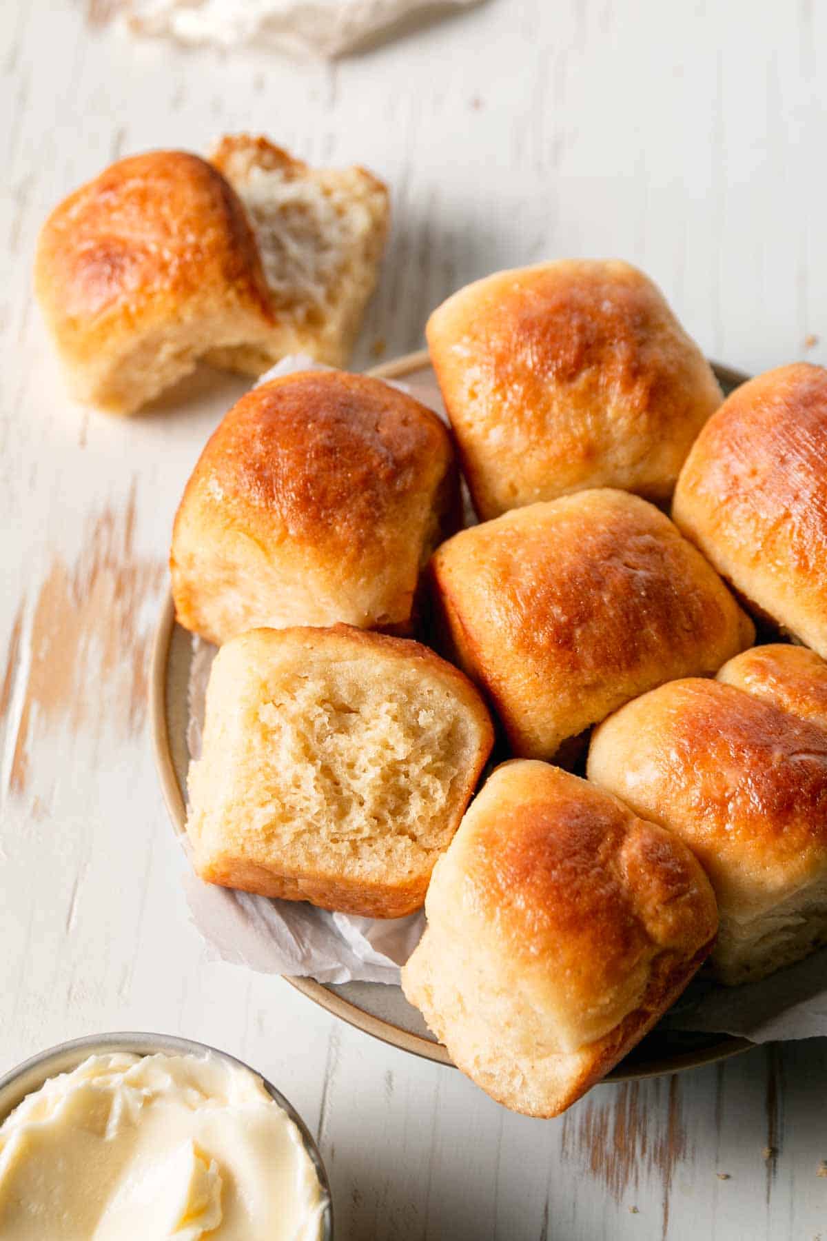 A plate filled with gluten-free hawaian rolls next to one split roll and a bowl of butter.