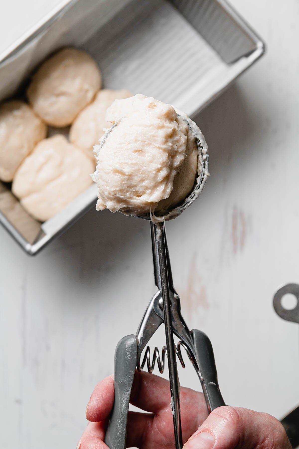 A cookie scoop holding sticky gluten-free roll dough above a pan of rolls.