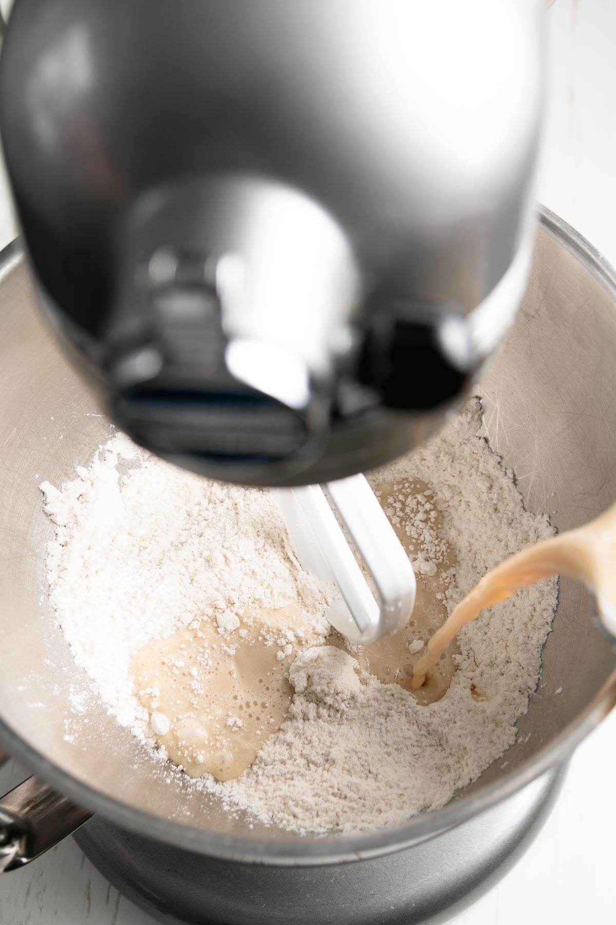 Pouring warm milk mixture into dry ingredients in a stand mixer bowl with a paddle attachment, ready to mix gluten-free Hawaiian roll dough.