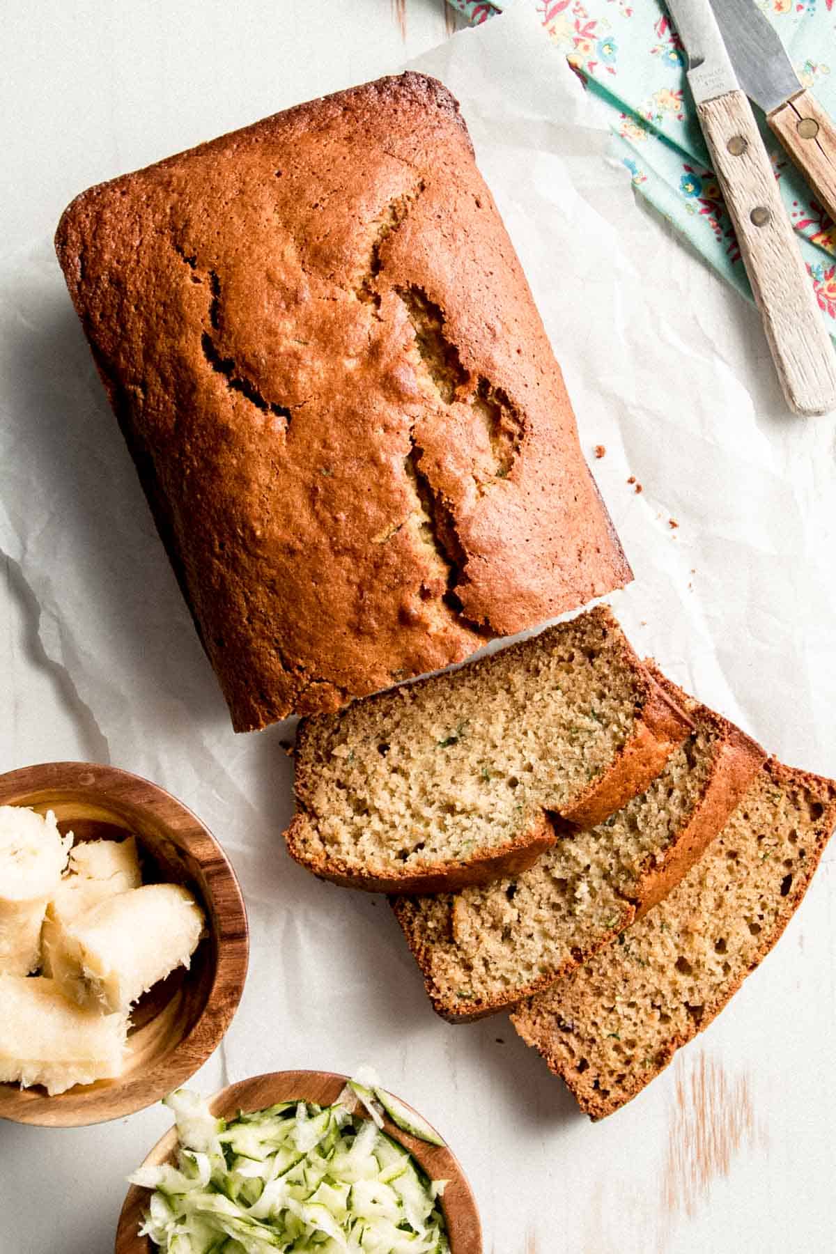 A loaf of Gluten-free Zucchini Banana Bread with a few slices laid on their sides next to small bowls of ripe banana and grated zucchini.