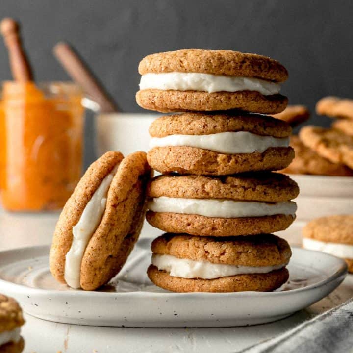 A stack of pumpkin cream pies, one cookie leaning against the stack. Pumpkin puree and a frosting bowl in the background.