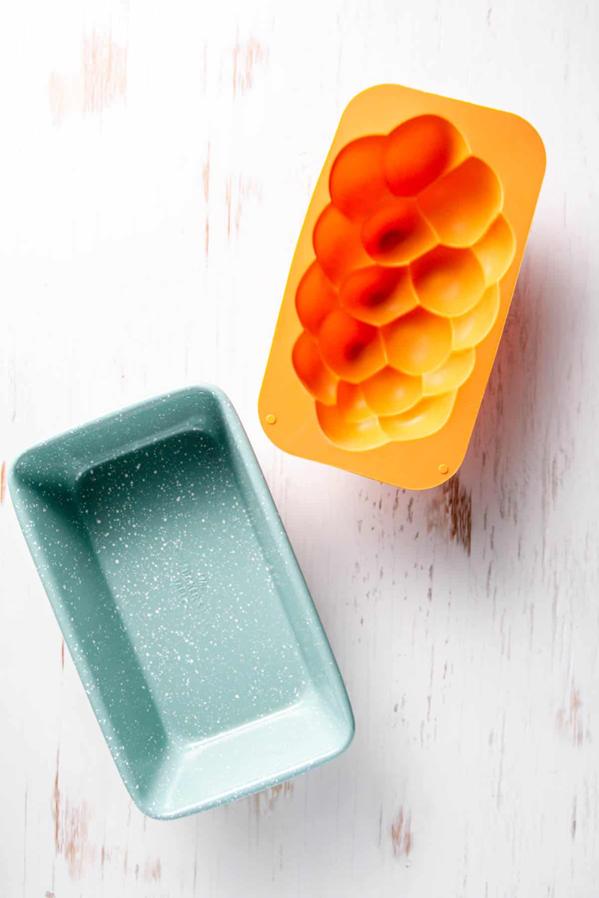 A standard size green speckled loaf pan and an orange silicone challah bread mold on white wooden board.