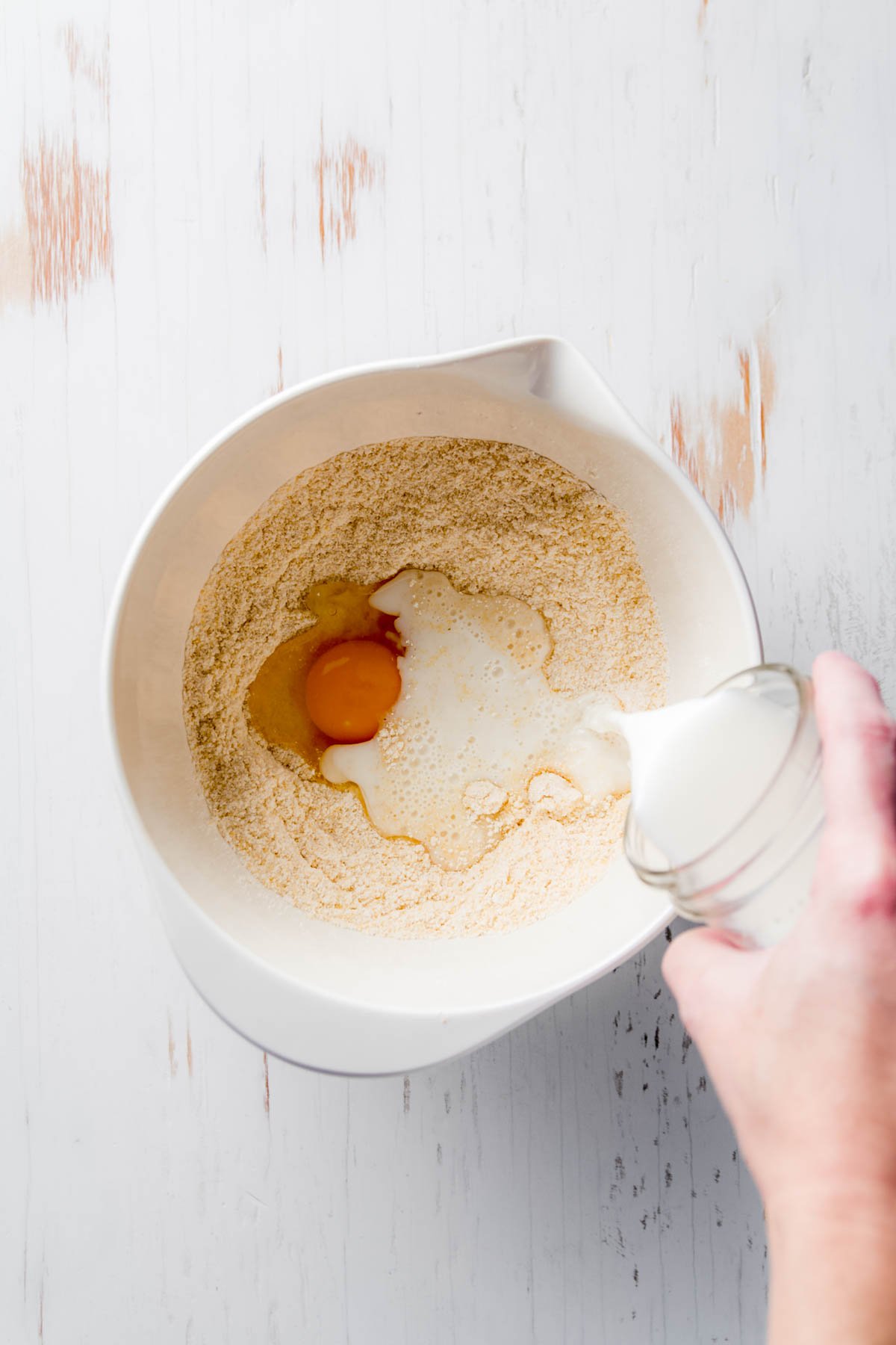 A white mixing bowl with cornbread batter being whisked until smooth. A hand holds the bowl steady while another hand stirs with a metal whisk.
