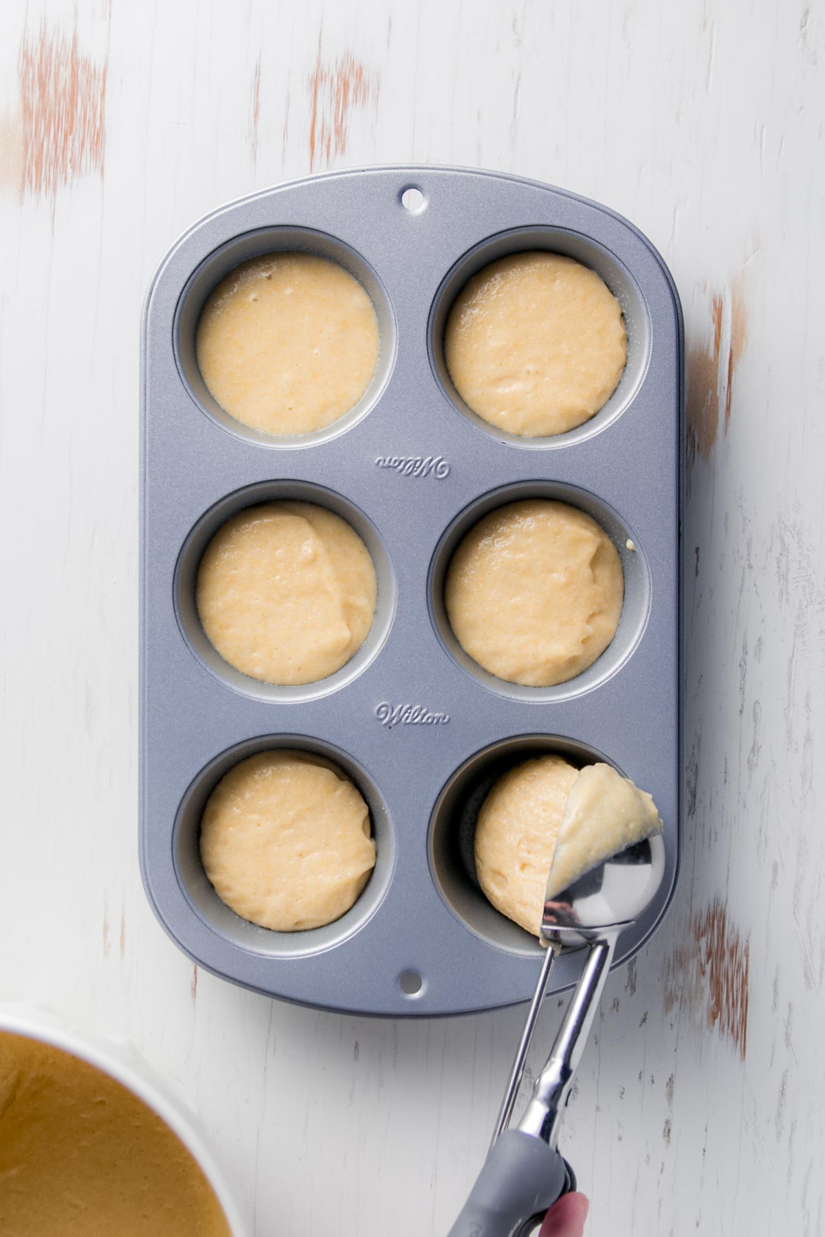 A metal muffin tin filled with cornbread batter. A hand is using a cookie scoop to portion the batter into the cups.