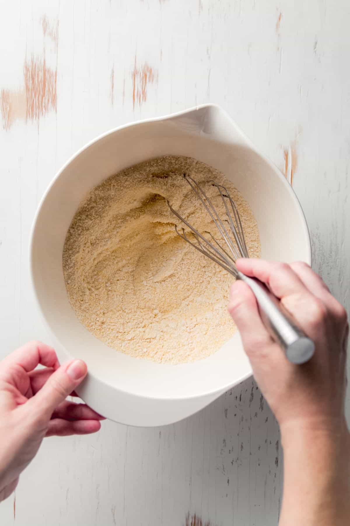 A white mixing bowl filled with dry cornbread mix, with an egg and milk being poured in from a small glass jar. The bowl sits on a white wooden surface.