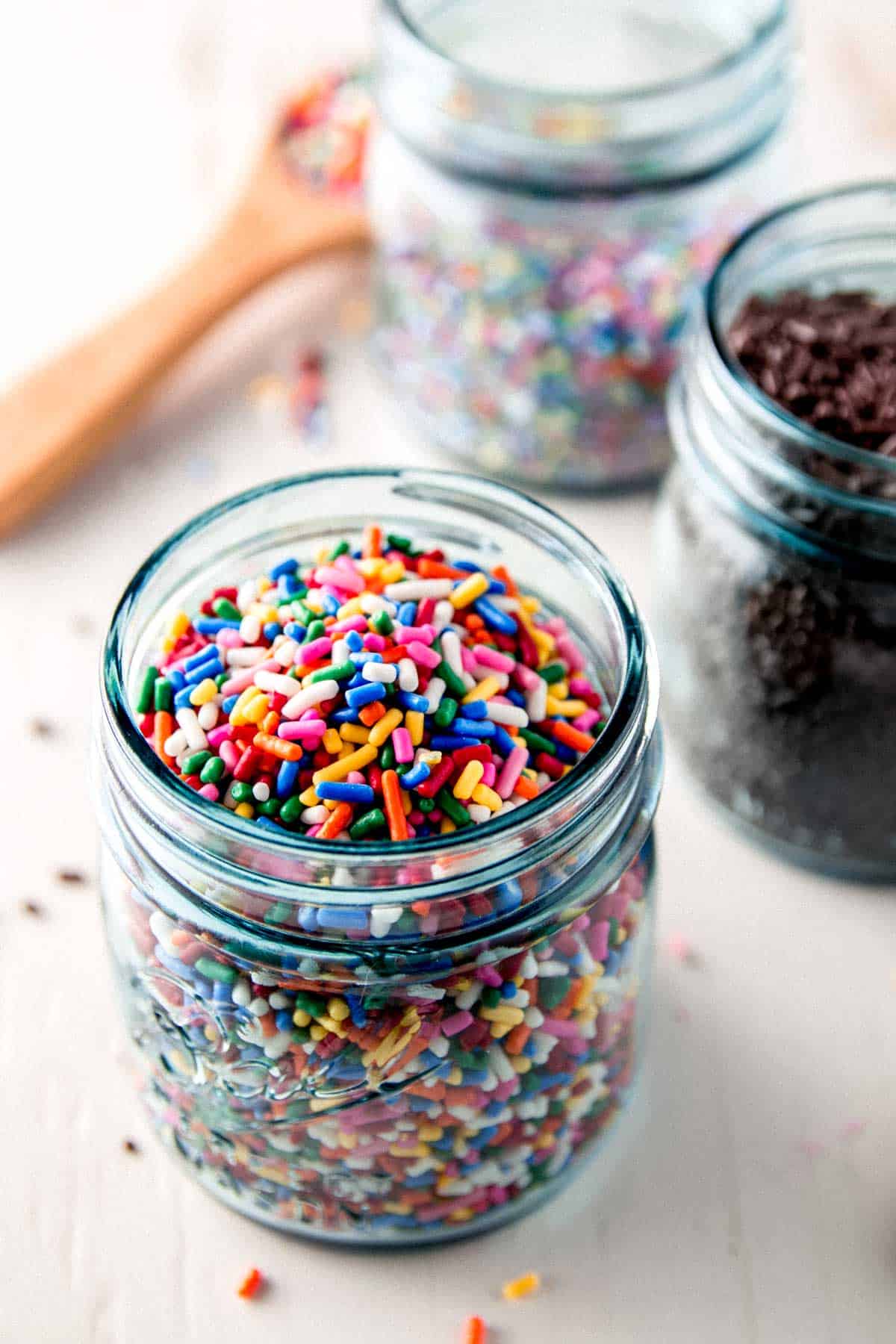 Three blue mason jars filled with gluten free sprinkles on white wooden board.