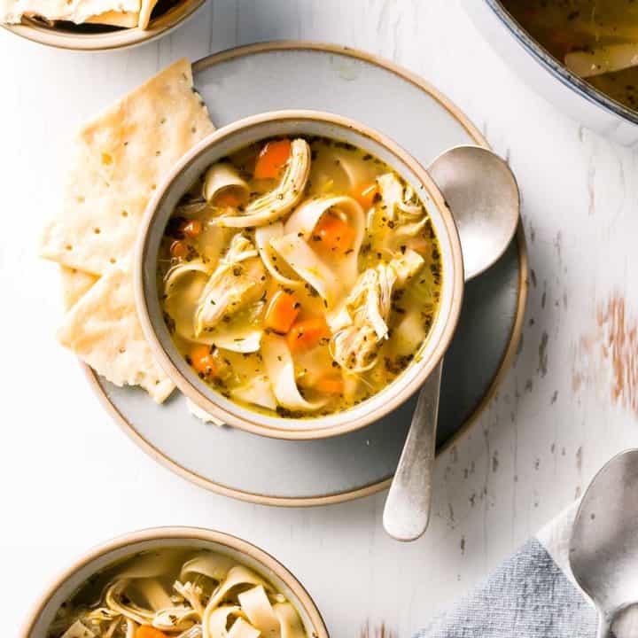A bowl of gluten free chicken noodle soup shown from above with crackers and a spoon to the side.