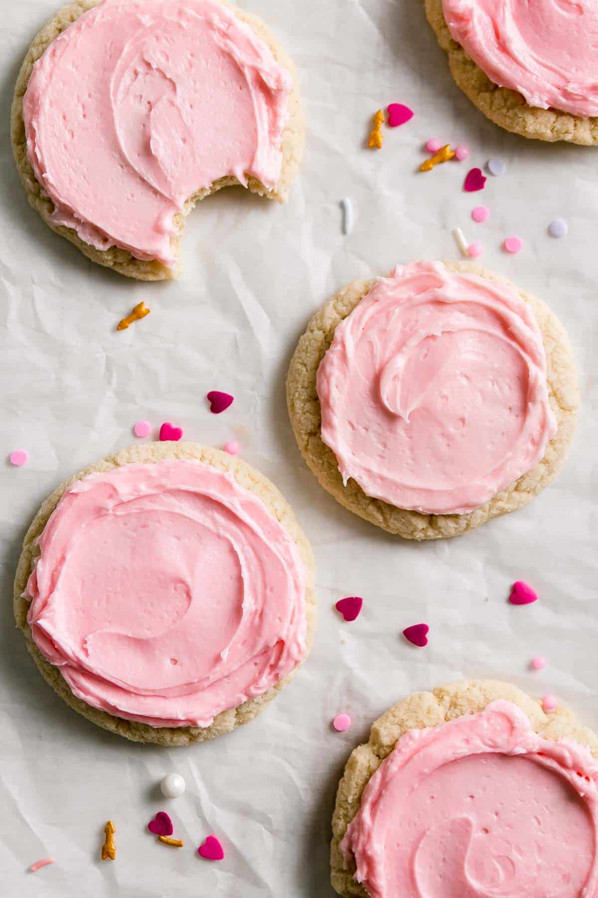 Sugar cookies topped with swirl of pink frosting scattered on crumpled parchment. Heart shaped sprinkles scattered around.