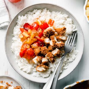 A shallow bowl filled with diced breaded chicken, red bell peppers, onion, and pineapple in sweet fire sauce atop a bed of rice.