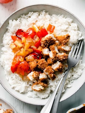A shallow bowl filled with diced breaded chicken, red bell peppers, onion, and pineapple in sweet fire sauce atop a bed of rice.