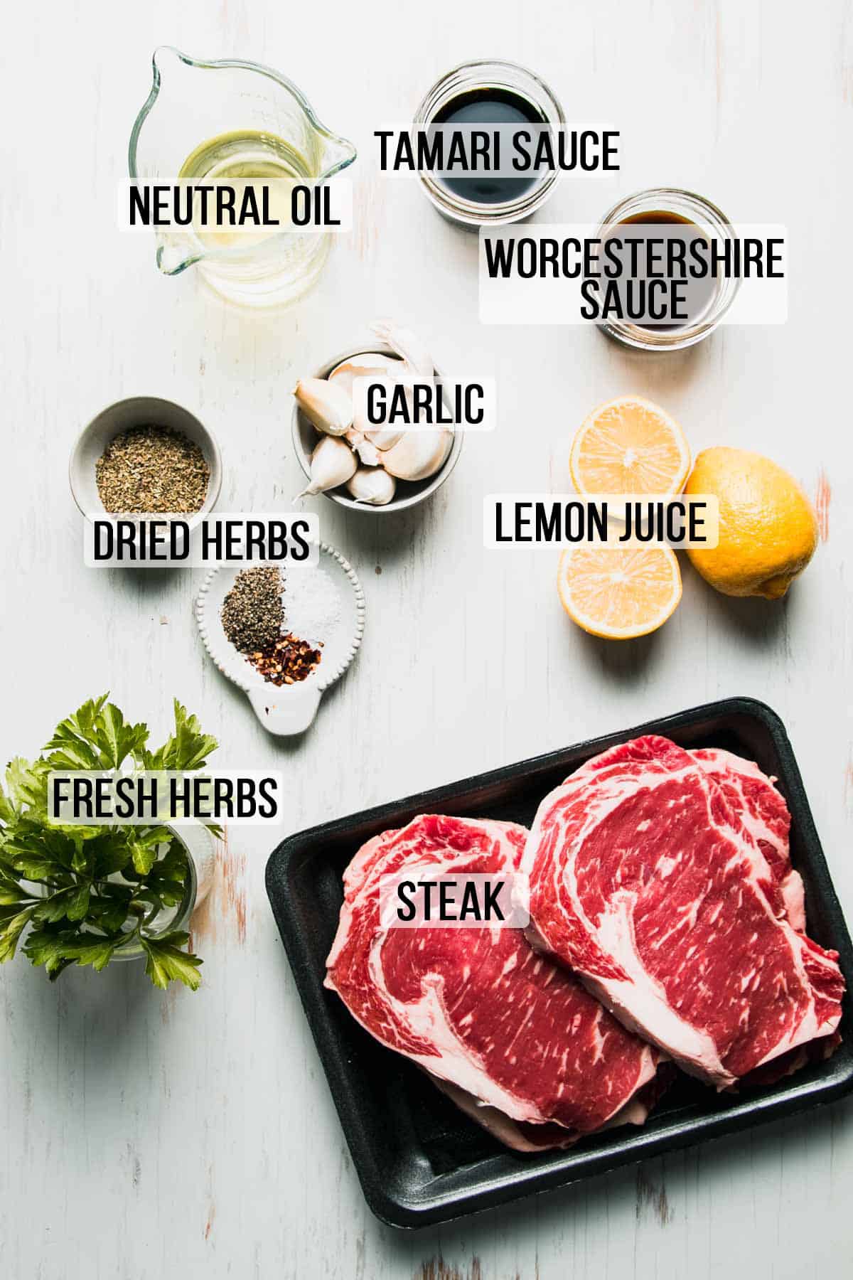 Ingredients for marinade and steak laid out on white wooden surface labeled with text, "neutral oil, Tamari sauce, Worcestershire sauce, garlic, dried herbs, lemon juice, fresh herbs, steak."